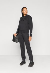 Μαύρο φούτερ με κορδόνια και τσέπη μπροστά, συνδυασμένο με μαύρο παντελόνι φόρμας. Το μοντέλο κρατά μια μαύρη τσάντα. Και τα δύο ρούχα είναι κατασκευασμένα από απαλό ύφασμα.