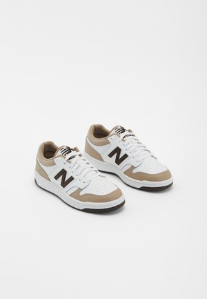 Ein Paar weiße und beige New Balance Sneakers mit schwarzem "N"-Logo, durchlöcherter Zehenpartie und weißen Schnürsenkeln auf einem schlichten weißen Hintergrund.