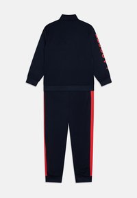 Ensemble de survêtement bleu marine foncé comprenant une veste zippée avec du texte rouge sur la manche et un pantalon assorti avec des rayures rouges sur les côtés. Texture de tissu lisse.