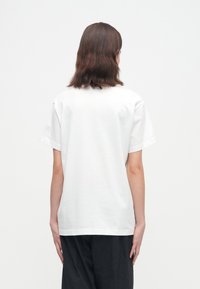 Vállig érő barna hajú személy egyszerű fehér rövidujjú pólót és fekete nadrágot visel, háttal áll a fehér háttér előtt.