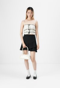 Svart volangskjørt, hvit bluse med svarte detaljer, hvit veske og svarte sko. Modellen har på seg hvite sokker og står på en ensfarget bakgrunn.