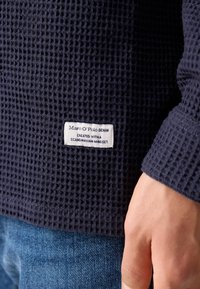 Tamsiai mėlynas vaflinis megztinis su etiketėle, ant kurios užrašyta "Marc O'Polo DENIM", prisiūta apačioje, derinama su mėlynos džinsinės kelnės.