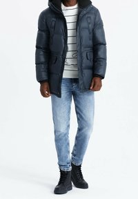 Giacca imbottita blu navy con cappuccio, due tasche frontali e dettagli con bottoni, abbinata a un maglione a righe e jeans blu, e stivali neri con lacci.