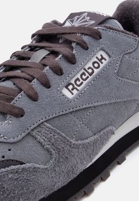 Γκρι σουέντ αθλητικό παπούτσι Reebok με καφέ κορδόνια, ορατό λογότυπο Reebok στο πλάι, υφασματικές λεπτομέρειες και μαύρη και λευκή σόλα.