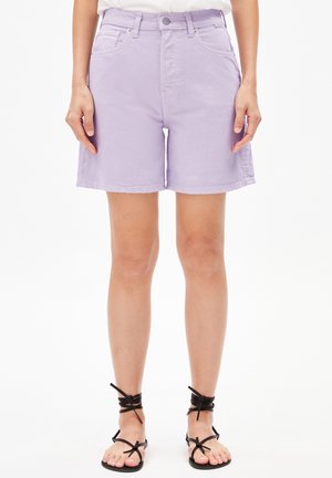 Mujer con pantalones cortos vaqueros de color lila claro y sandalias negras con tiras, de pie sobre un fondo blanco liso.