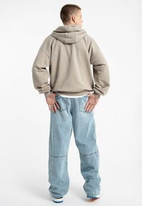 Beżowa bluza z dużym kapturem, luźny krój. Jasnoniebieskie workowate dżinsy z szwami i widocznymi kieszeniami. Gładka faktura, minimalne detale.