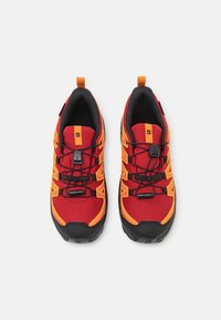 Scarpe da ginnastica rosse e nere con accenti arancioni, tomaia in rete, suola in gomma, lacci elastici e un manico sul tallone.