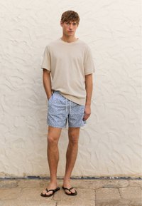 Camicetta beige a maniche corte, pantaloni da bagno blu a fantasia con accenti bianchi e infradito nere; in piedi contro un muro bianco strutturato.