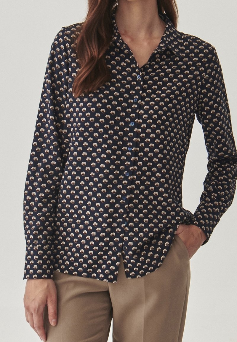 Femme portant une chemise bleu marine à manches longues avec de petits motifs floraux blancs et un pantalon beige, une main dans la poche.