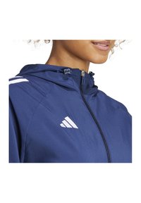 Giacca sportiva blu navy con chiusura a zip e cappuccio regolabile, caratterizzata da logo bianco e strisce di accentuazione sulle spalle. Texture liscia.