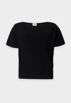 Femme debout portant un haut noir à manches courtes, un jean gris large et court, et des bottines noires à talons pointus.
