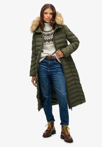 Groene lange pufferjas met een bontomranding op de capuchon, gecombineerd met een gebreid patroontrui, blauwe jeans en bruine veterschoenen. Geweven, gestikte ontwerpen.