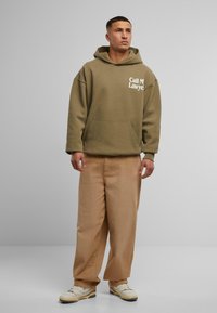 Olivenfarget oversized hettegenser med frontlomme og hvit tekst "Call My Lawyer." Kombinert med lysebeige bukser med løs passform og hvite sneakers.