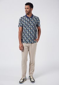 Marinblå mönstrad pikétröja med blå och guldblad design, i kombination med ljusbeige byxor och svarta sneakers med vita accenter.