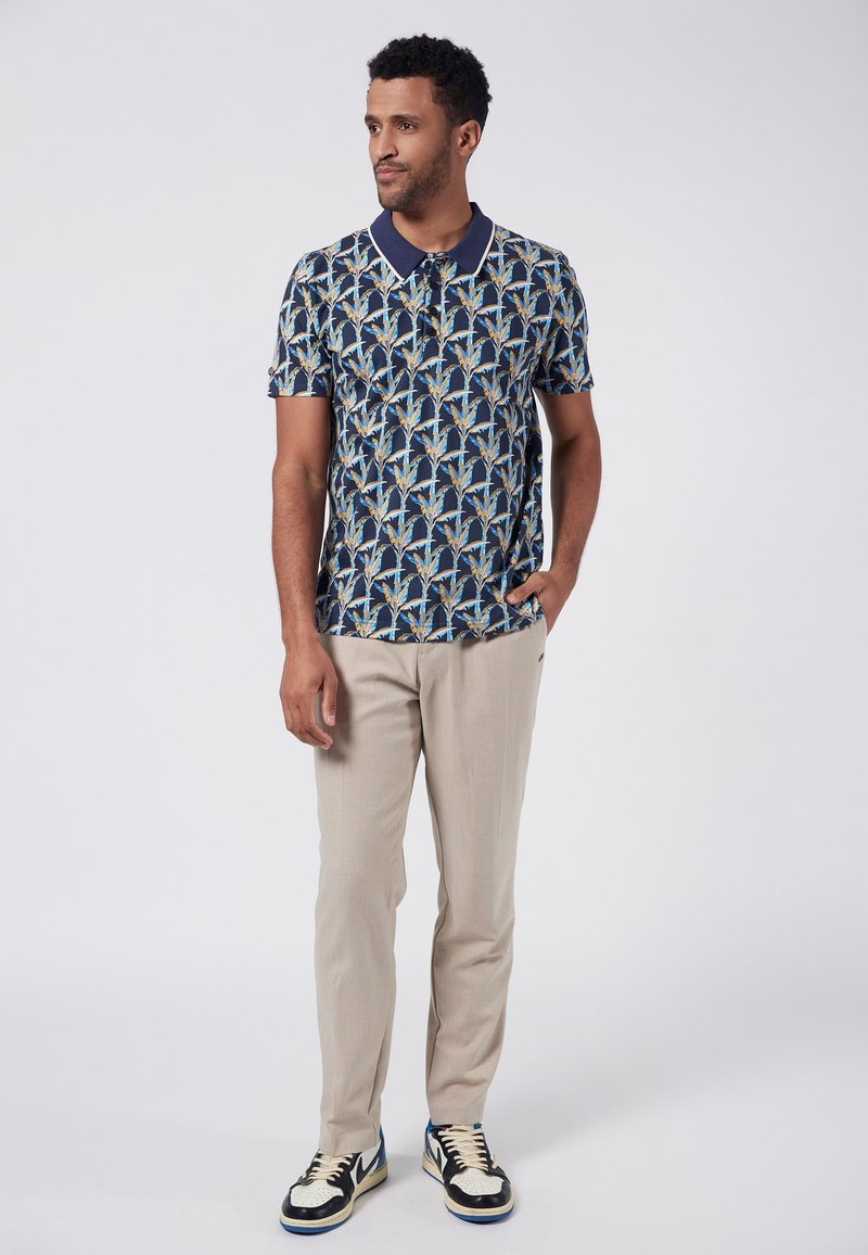 Marinblå mönstrad pikétröja med blå och guldblad design, i kombination med ljusbeige byxor och svarta sneakers med vita accenter.