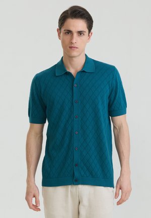 Ung mand iført en teal kortærmet skjorte med knapper og diamantmønster, stående mod en ensartet lys baggrund.