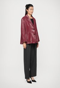 Blazer in ecopelle borgogna con design strutturato, ampi rever e due tasche frontali, abbinato a pantaloni a gamba larga con righe verticali neri e tacchi.