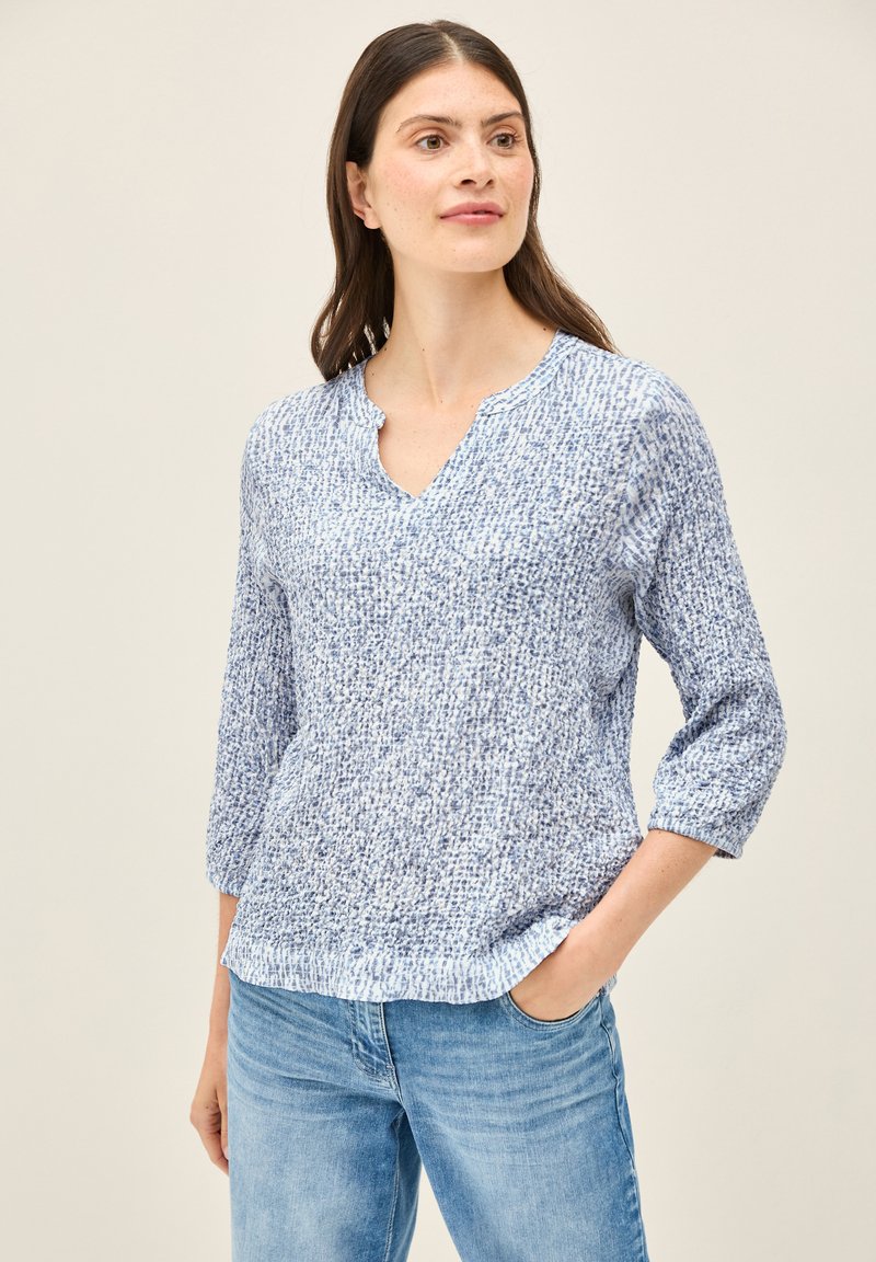 Haut en tricot texturé bleu doux avec un décolleté en V et des manches trois-quarts, présentant un motif subtil. Associé à un jean en denim bleu clair.