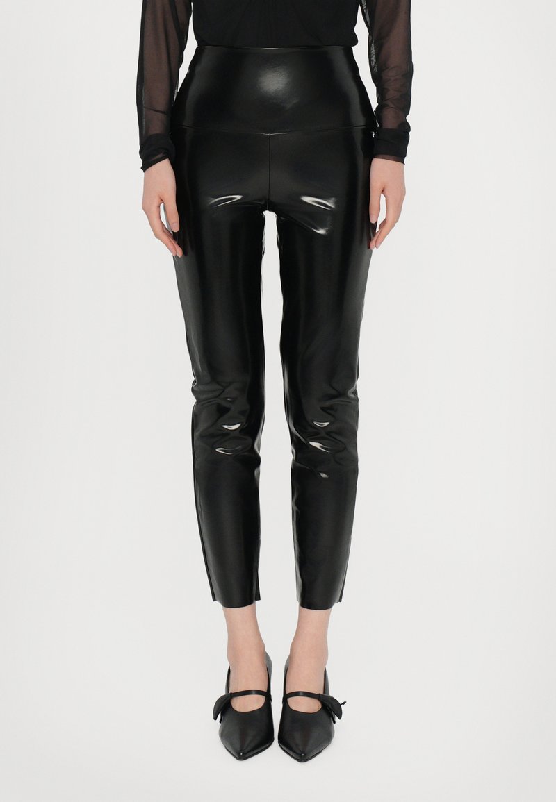 Højtaljede sorte leggings i imiteret læder med en blank overflade, tætsiddende pasform og ankellængde. Båret med sorte spidse flade sko med sløjfer.