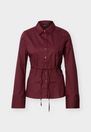 Burgunderfarget skjorte med lange ermer, knapper og krage, med tynt matchende belte knyttet i midjen, vist på en enkel lys bakgrunn.