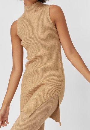 Haut long beige en maille côtelée sans manches avec fente latérale, porté avec un pantalon côtelé assorti sur une personne les bras légèrement étendus.