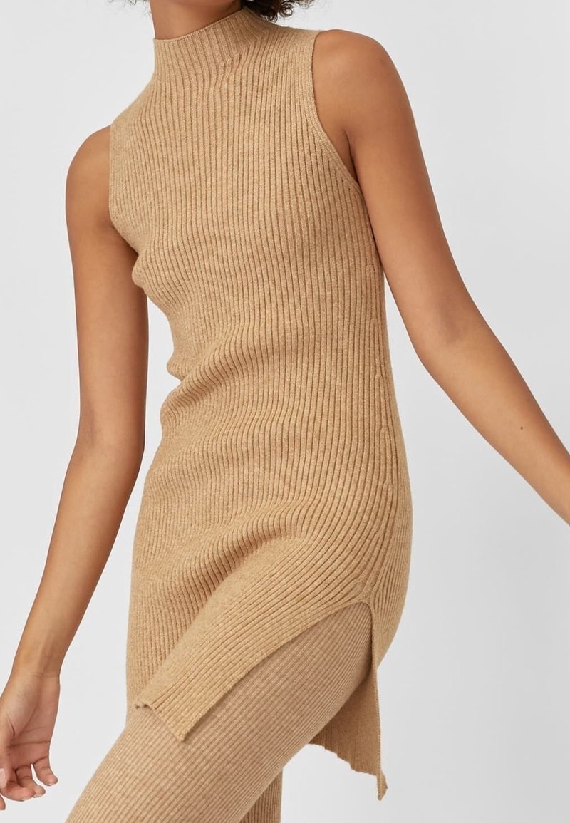 Haut long beige en maille côtelée sans manches avec fente latérale, porté avec un pantalon côtelé assorti sur une personne les bras légèrement étendus.