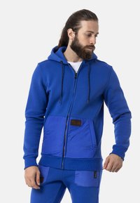 Blauer Reißverschluss-Hoodie aus strukturiertem Stoff, mit einer kontrastierenden blauen Fronttasche und einem kleinen Logo-Patch. Verstellbare Kapuze mit Kordelzug.