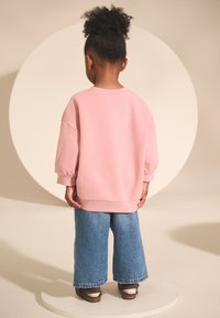 Felpa rosa oversize con polsini a coste, abbinata a jeans di denim blu a gamba larga. Lo sfondo presenta un elemento decorativo circolare.