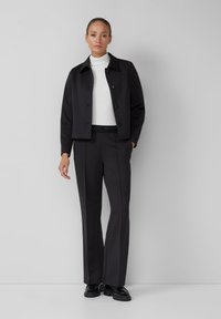 Veste noire courte avec des boutons, accompagnée de pantalons noirs ajustés dotés d'une taille texturée. Le modèle porte un pull col roulé blanc et des chaussures noires.