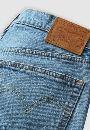 Blue denim jeans featuring a brown leather patch with embossed logo, contrasting orange stitching, and defined back pocket design.