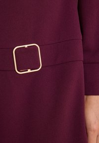 Close-up of a dark purple fabric dress with a square beige buckle on the waist belt, showing part of a person's hand.