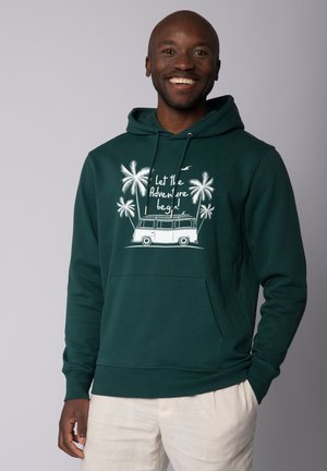 Donkergroene hoodie met een kangoeroezak, voorzien van een witte afbeelding van een bus en palmplanten met de tekst "Laat het avontuur beginnen!"