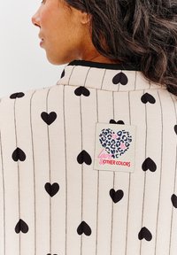 Svetlo béžový odev s čiernymi vzormi srdca a vertikálnymi pruhmi. Textilná visačka zobrazuje dizajn srdca s textom "láska a iné farby."