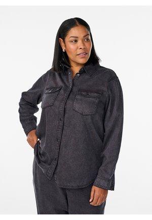 Woman with medium skin and straight black hair wearing dark gray buttoned shirt and matching pants, looking right with hand in pocket.