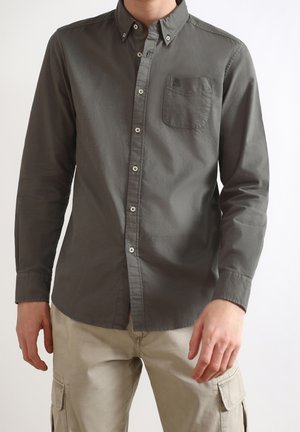 Mężczyzna ubrany w ciemnoszarą koszulę z długim rękawem zapinaną na guziki z kieszenią na piersi oraz beżowe bojówki na tle jednolitego tła.