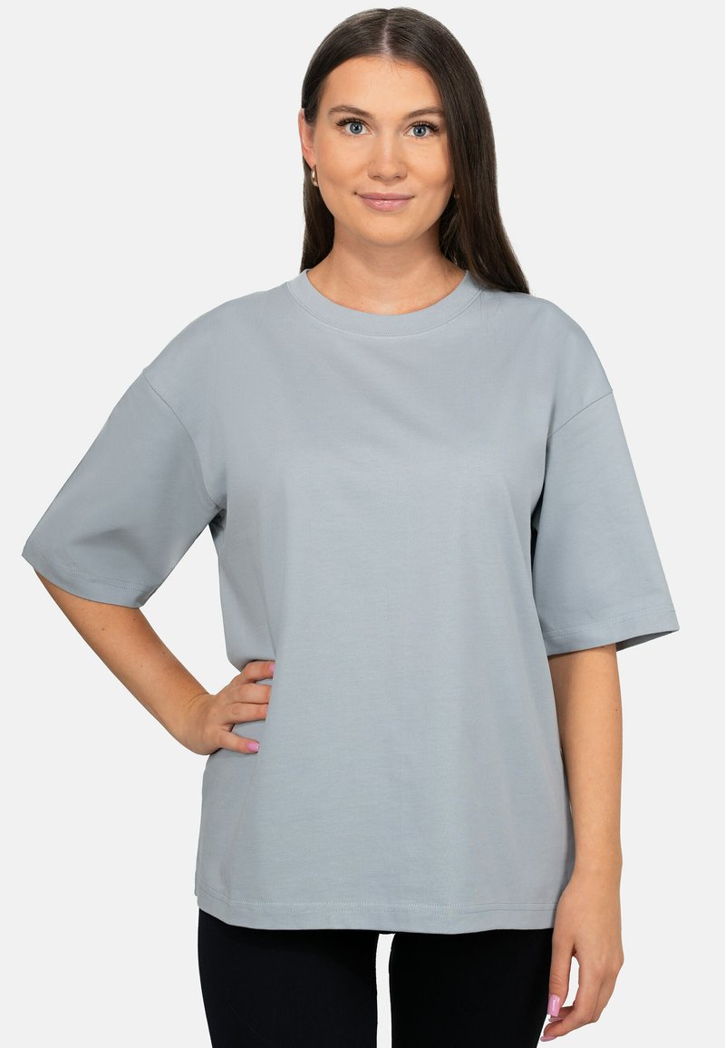 Donna con i capelli castani lunghi indossa una t-shirt corta a maniche corte grigio chiaro oversize e pantaloni neri, in piedi con una mano sul fianco.