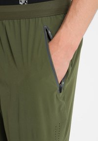 Pantaloni verde oliva con tasca con zip, vita elastica e tessuto traspirante. Texture sottile con fori di ventilazione vicino alla tasca.