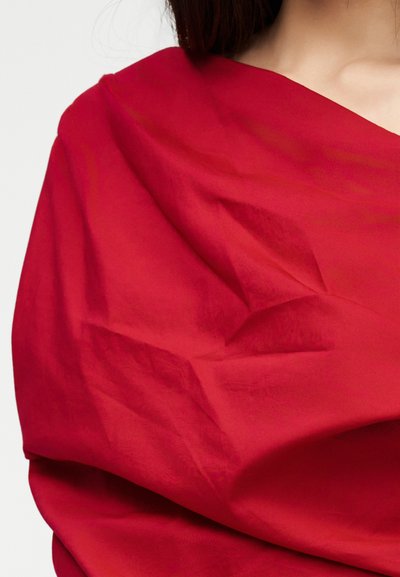 Γυναίκα με φωτεινό κόκκινο ύφασμα που πέφτει ασύμμετρα πάνω στον κορμό και τον ώμο της, σε απλό φόντο.