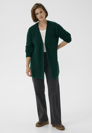 Cardigan tricotat de culoare verde închis, cu o croială lejeră, deschis în față și cu mâneci lungi, combinat cu pantaloni negri cu picior drept și pantofi cu talpă înaltă maro.