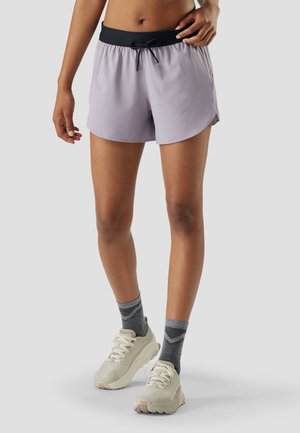 Kvinde iført lyse lilla træningsshorts med sort taljebånd, grå ankelsokker og off-white løbesko foran en ensfarvet baggrund.