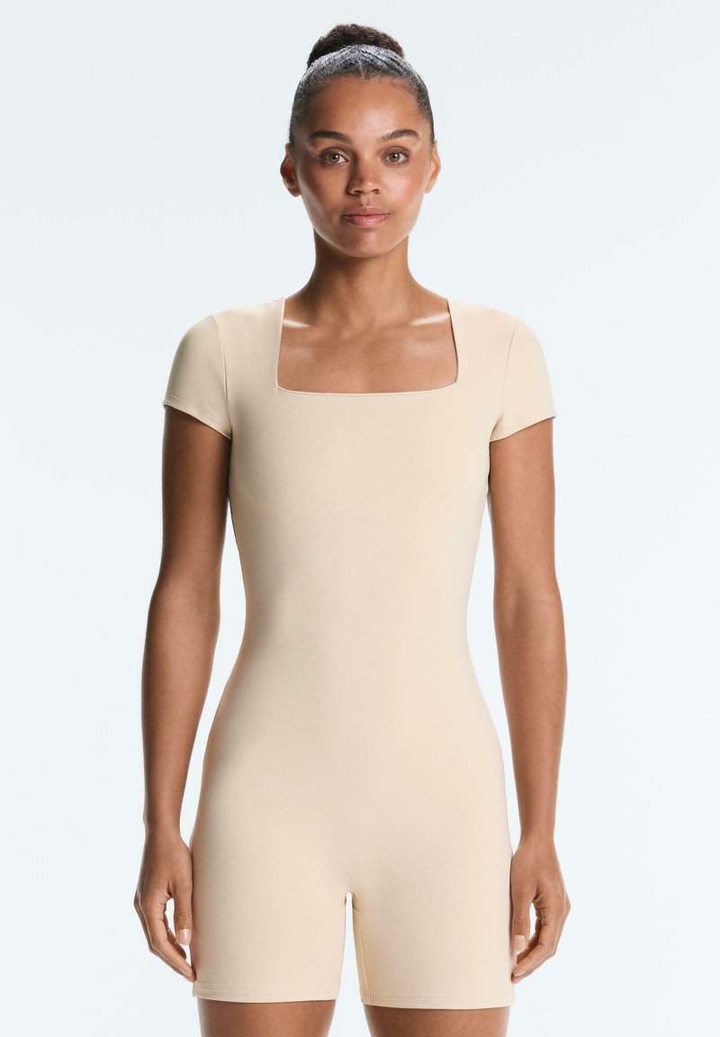 Femme avec les cheveux en chignon portant une combinaison beige ajustée à manches courtes et col carré, debout devant un fond clair uni.