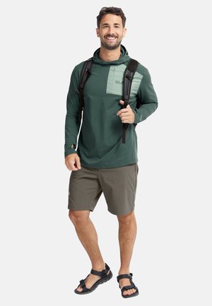 Lächelnder Mann, der einen grünen Langarm-Hoodie, khakifarbene Shorts, schwarze Wandersandalen und einen schwarzen Rucksack trägt, steht vor einem weißen Hintergrund.