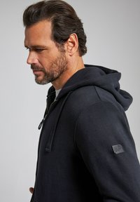 Schwarze Zip-Hoodie mit Kordelzugkapuze, aus einem weichen Material gefertigt, mit einem Logo-Patch am Ärmel und einer Kängurutasche.