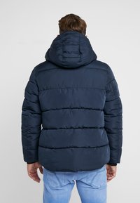 Marineblauwe pufferjas met capuchon, uitgevoerd met een doorgestikt ontwerp en een gestructureerde stof. Mouwboorden en zoom zijn afgewerkt met elastische banden.