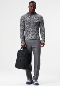 Gestrickter grauer und schwarzer Pullover mit langen Ärmeln, graue maßgeschneiderte Hose und einen schwarzen Rucksack, der in einer Hand gehalten wird. Die Sneaker haben graue und blaue Akzente.