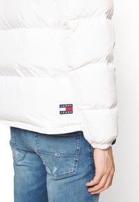 Hvid dynejakke med quiltet design, elastiske manchetter og et lille mærke med logo. Bæres over blå slidte jeans. Blød, glat tekstur.