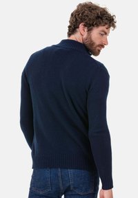 Pull en maille bleu marine avec un col montant, des poignets et un ourlet côtelés. Texture lisse, coupe ajustée, vu de dos. Un jean est visible.