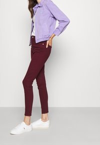 Chaqueta de mezclilla morada con cremallera en el frontal, combinada con pantalones ajustados burdeos y zapatillas blancas. Presenta un logo bordado en la chaqueta.