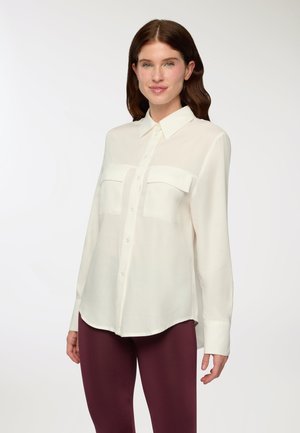 Camicia button-up color crema con colletto a punta, due tasche sul petto e maniche lunghe, abbinata a leggings bordeaux. Texture liscia.