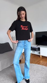 Femme ajustant un t-shirt noir « Sorry Tokyo », portant un jean bleu clair et des chaussures noires, debout dans un salon avec TV et canapé.
